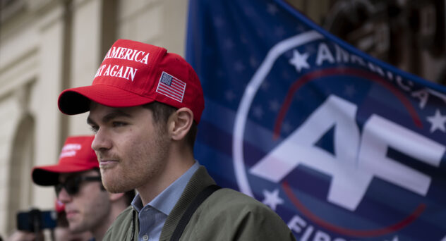 Nick Fuentes wearing a MAGA hat, standing in front of an America First flag.