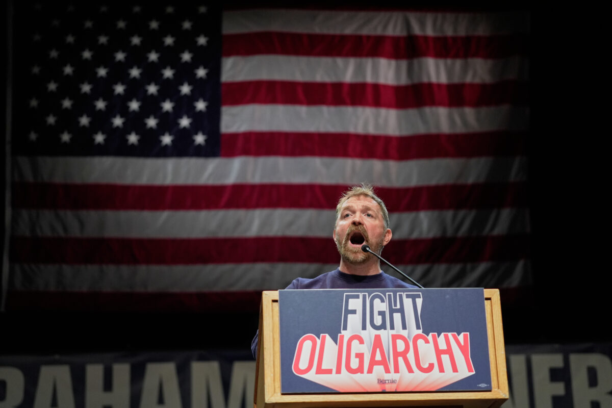 Graham platner at a podium with an american flag behind him. the podium has "fight oligarchy" onit