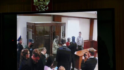 photo of a screen showing stephen hubbard standing in a glass box in a courtroom in russia surrounded by people