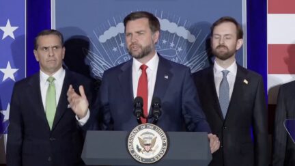 Vance speaking at a podium with two men behind him